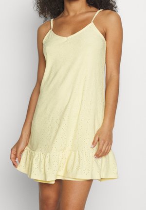 Žena v bezrukávových bledě žlutých šatech s dírkovaným vzorem, tenkými ramínky a volánkovým lemem, stojící před jednobarevným pozadím.
