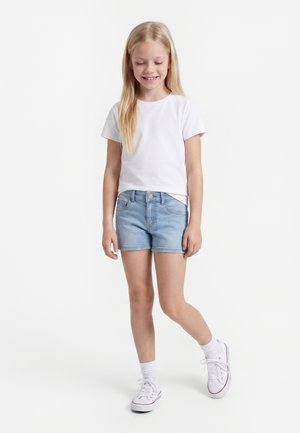 Młoda dziewczyna uśmiecha się, ubrana w białą koszulkę, jasnoniebieskie szorty dżinsowe, białe skarpetki i białe trampki, stoi na białym tle.