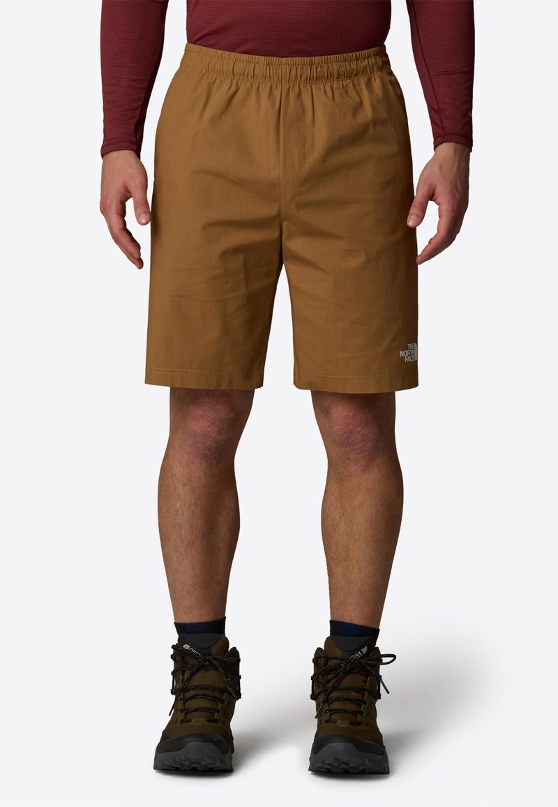 Pantalones cortos marrones hechos de un material ligero, con una cintura elástica y un pequeño logotipo en la parte inferior de la pierna. Llevados con botas de senderismo.