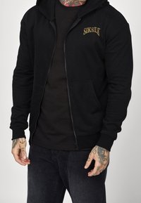 Sudadera con cremallera negra hecha de tejido suave, con un bolsillo frontal y un logo bordado. Incluye mangas largas y capucha.
