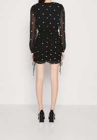 Svart klänning med genomskinliga ärmar, vita prickar, rynkad design, dragsko-detaljer och knälång nederdel. I kombination med glansiga svarta klackar.