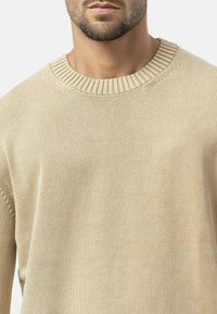 Beige gestrickter Pullover mit rundem Rippkragen, der eine glatte Textur und subtile Details an den Schultern aufweist.