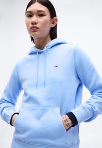 Νεαρός/Νεαρή που φοράει ανοιχτό μπλε φούτερ με μικρό λογότυπο, τα χέρια στην μπροστινή τσέπη, δείχνοντας τατουάζ στον καρπό, σε απλό λευκό φόντο.