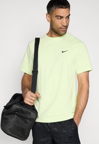 Ανοιχτό πράσινο μπλουζάκι Nike με κοντά μανίκια και μαύρο λογότυπο, συνδυασμένο με μια υφασμάτινη μαύρη τσάντα με φερμουάρ και ρυθμιζόμενο ιμάντα.