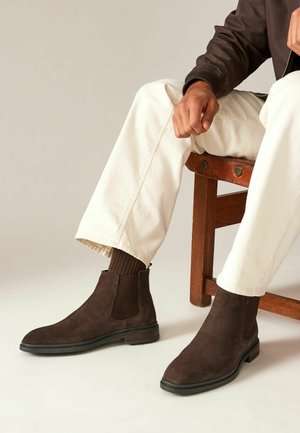 Botas de tobillo de ante marrón con paneles elásticos en los laterales, puntera redondeada y suela de goma negra. Combinadas con pantalones crema y calcetines de canalé marrones.