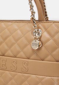 Brązowa pikowana skórzana torebka na łańcuszku z okrągłymi złotymi zawieszkami marki Guess z napisem "GUESS EST 1981".