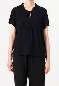 Blouse noire à manches courtes avec un col à volants et un détail à nouer; tissu texturé avec de petits points en relief et une coupe décontractée et fluide.