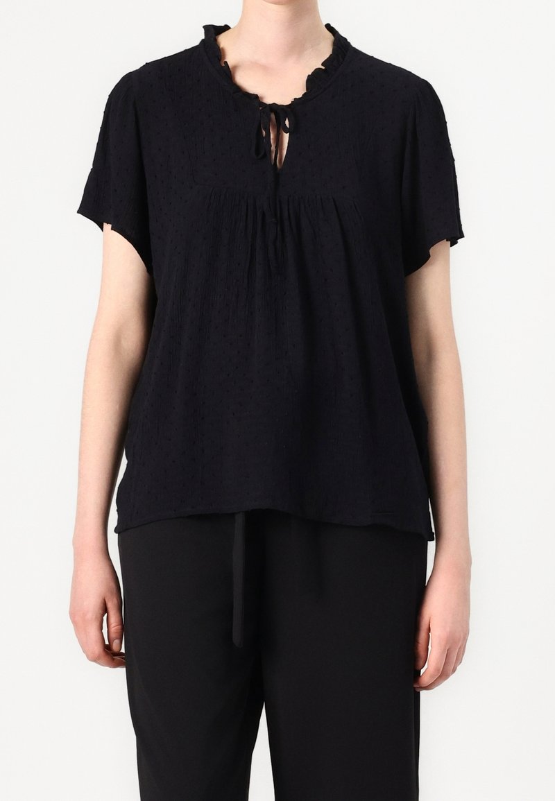 Blouse noire à manches courtes avec un col à volants et un détail à nouer; tissu texturé avec de petits points en relief et une coupe décontractée et fluide.