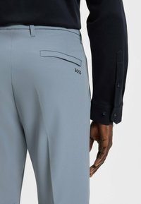 Light gray trousers made of smooth fabric, featuring a back pocket and the "BOSS" logo in dark lettering, paired with a dark buttoned shirt.