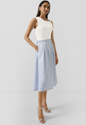 Femme aux longs cheveux noirs portant un haut blanc sans manches, une jupe plissée midi bleu clair et des sandales argentées à talons hauts, debout devant un fond uni.