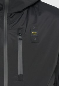 Schwarze Jacke mit einer wasserdichten Reißverschlusstasche, einem Logo-Patch und einer glatten Stoffstruktur. Das Design umfasst minimalistische Hardware-Akzente.