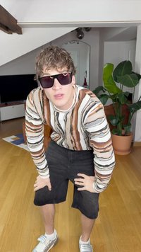 Pikkade varrukatega mustriga särk pruunide, oranžide ja mustade triipudega, mustad lühikesed püksid ja prillid. Seisab puidust põranda peal, siseruumis.