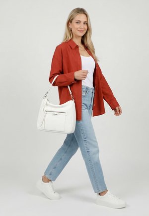 Jonge vrouw die loopt, draagt een rood shirt, wit topje, lichtblauwe spijkerbroek, witte sneakers en draagt een witte handtas over haar schouder.