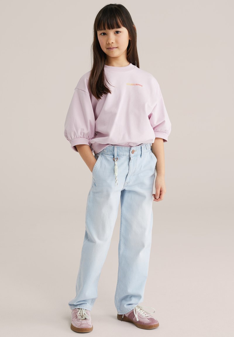 Jeune fille aux cheveux longs et foncés portant un sweat-shirt à manches bouffantes violet clair, un jean bleu clair avec porte-clés et des baskets violettes.