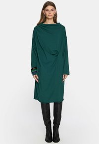 Kobieta ubrana w długą, luźną ciemnozieloną sukienkę z wysokim dekoltem, w połączeniu z czarnymi kozakami do kolan i dużymi kolczykami w kształcie kół.