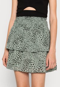 Femme portant une mini-jupe à volants de couleur vert clair avec un motif de points noirs irréguliers et un haut noir court avec une découpe à l'avant.