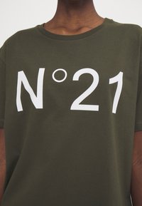 Camiseta de algodón verde oliva con cuello redondo que presenta un gran texto impreso en blanco "N°21" en el frente. Textura suave y ajuste relajado.