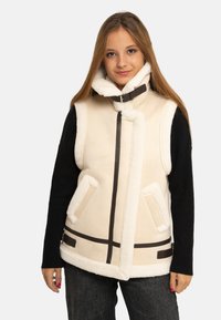 Jeune femme portant un gilet sans manches en peau lainée crème avec col à boucle, par-dessus un pull noir à manches longues et un jean foncé, debout devant un fond blanc.