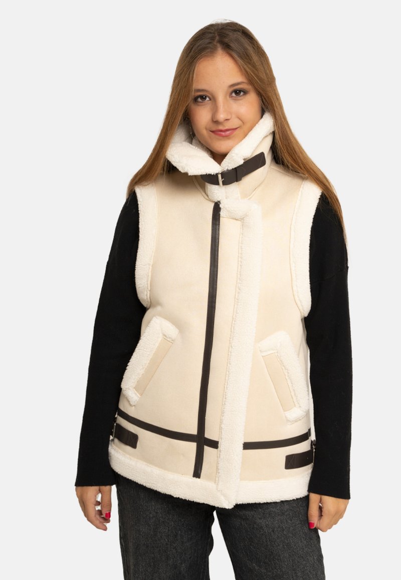 Jeune femme portant un gilet sans manches en peau lainée crème avec col à boucle, par-dessus un pull noir à manches longues et un jean foncé, debout devant un fond blanc.