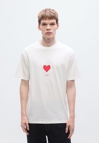 Λευκό βαμβακερό μπλουζάκι με γραφικό κόκκινης καρδιάς οριγκάμι και το κείμενο «HUGO» από κάτω. Κοντά μανίκια και άνετη εφαρμογή.