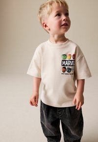 Kremno bele kratke majice s prikazom glav likov iz Marvela in besedilom, združene s temno sivimi širokimi kavbojkami. Tekstura se zdi mehka.