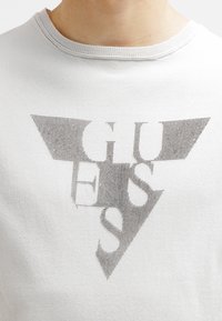 Biała bawełniana koszulka z dużym szarym nadrukiem logo "GUESS" w trójkowym wzorze, o teksturowanym, kropkowanym wyglądzie.