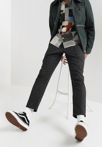 Grüne Zip-Jacke, gemustertes kurzärmliges Hemd, gestreifte Hose, schwarz-weiße Sneaker mit strukturierten Sohlen, weiße Socken.