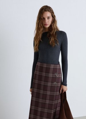 Woman with long wavy hair wearing a dark ribbed sweater and a brown plaid skirt with metal ring details, holding a brown coat.