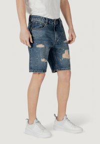Shorts di jeans con un design distressato, strappi sfilacciati, lavaggio chiaro e leggeri schizzi di vernice, abbinati a sneakers bianche.