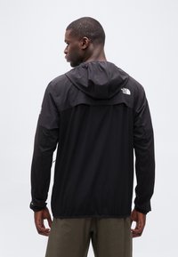 Άνδρας που φοράει μαύρο μπουφάν με κουκούλα και το λογότυπο της The North Face στην πλάτη και το μανίκι, στέκεται με την πλάτη προς το φακό σε ουδέτερο φόντο.