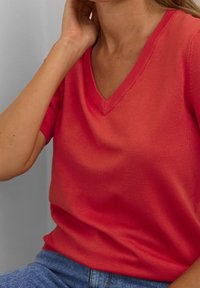 V-neck coral knit top with short sleeves, smooth texture, and a fitted design. Paired with blue denim jeans.