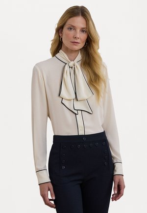 Blusa crema con colletto a fiocco annodato, piping nero lungo i bordi, maniche lunghe e bottoni sul davanti, abbinata a pantaloni scuri a vita alta.