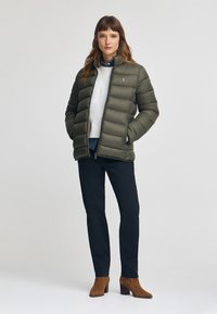 Olivgrön pufferjacka med horisontell quiltning, hög krage och dragkedja. Bärs över en vit sweatshirt och mörka jeans med bruna klackstövlar.