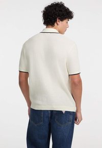 Crem farbenes Poloshirt mit kurzen Ärmeln aus strukturiertem Stoff, mit einem schwarz eingefassten Kragen, getragen zu dunkelblauen weit geschnittenen Jeans.