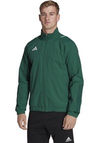 Grüne Sportjacke aus leichtem Material. Verfügt über einen durchgehenden Reißverschluss, elastische Bündchen und drei weiße Streifen an den Ärmeln.