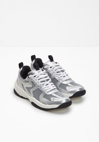 Sneaker in rete argento e nero con lacci bianchi, suole bianche spesse e tab di chiusura neri su uno sfondo bianco.
