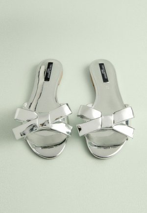 Stříbrné skluzné sandály s lesklým povrchem, zdobené širokou dekorativní mašlí a otevřenou špičkou. Plackové podrážky s hladkými okraji.