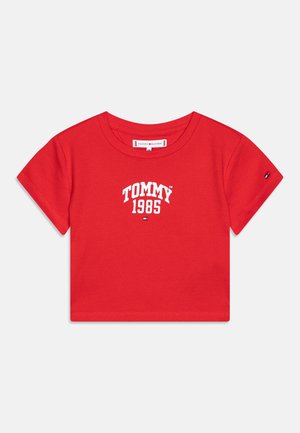 Camiseta de algodón roja con mangas cortas, que presenta el texto blanco "TOMMY 1985" centrado en el frente y un pequeño logo en la manga.