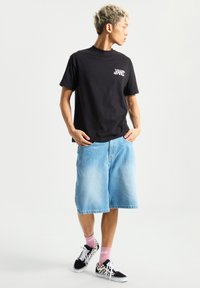 Sort kortærmet t-shirt, blå denim shorts knælang, lyserøde sokker og sort/hvide ternede sneakers. Enkel og afslappet design.