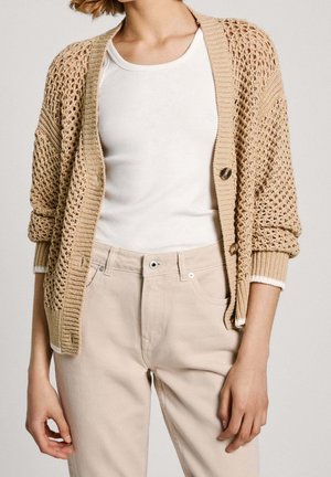 Personne portant un cardigan beige en maille ouverte sur une chemise blanche et un pantalon beige clair, debout, les bras détendus le long du corps.