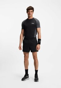 Zwarte sport t-shirt met grijze accenten en logo, gecombineerd met zwarte shorts. Accessoires omvatten een zwart polsbandje met logo. Hardloopschoenen zichtbaar.