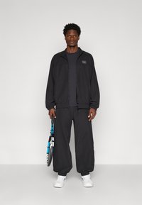 Veste de sport noire zippée avec des poignets côtelés, associée à un survêtement noir assorti. La personne tient une raquette de tennis avec des accents bleus.