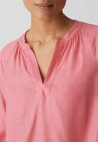 Pull en rose avec un col en V et un tissu doux. Se caractérise par une coupe décontractée et des fronces subtiles au niveau du col pour plus de texture.