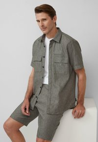 Lichtgrijs shirt met korte mouwen, twee borstzakken, donkere knopen en bijpassende shorts, gemaakt van een textuurstof, gedragen over een wit T-shirt.