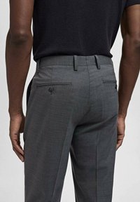 Pantaloni gri croiți, cu o textură netedă, două buzunare la spate cu nasturi și un design frontal plat. Inclusiv lauri pentru curea.