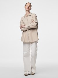 Smėlio spalvos marškiniai su sagomis, vertikaliomis juostelėmis, ilgomis rankovėmis ir vienu krūtinės kišeniu; derinami su baltomis platėjančiomis kelnėmis ir kasdieniais batais.