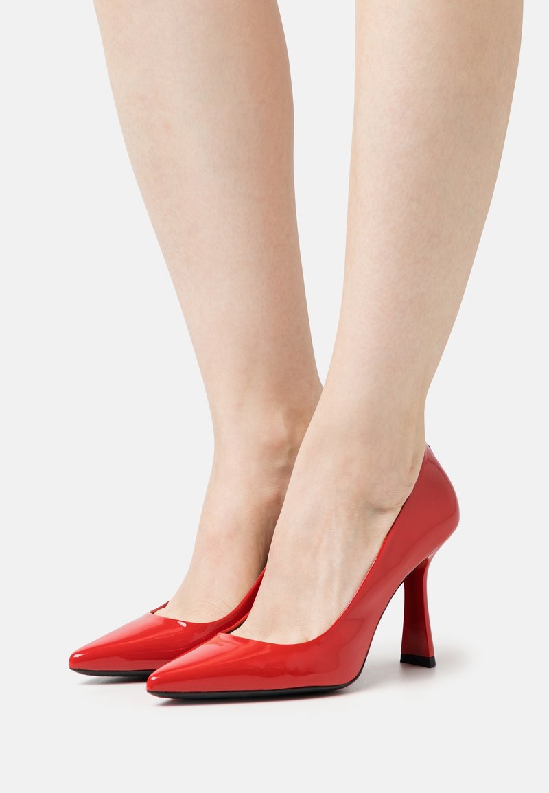 Jambes portant des escarpins rouges brillants à bout pointu avec un talon fin et courbé sur un fond clair uni.