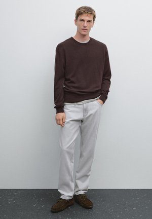 Brauner Strickpullover, graues T-Shirt darunter, hellgraue Hose und braune Schuhe. Das Model steht mit einer Hand in der Tasche vor einem schlichten Hintergrund.