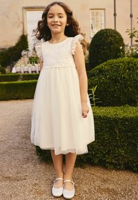 Witte jurk met geknoopte mouwen, geborduurde lijfje en gelaagde tule rok. Gecombineerd met witte Mary Jane schoenen, staand in een tuin.