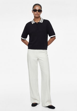 Femme portant un polo noir à manches courtes avec bordure blanche, pantalon large blanc, ballerines noires et lunettes de soleil noires, debout les mains derrière le dos.